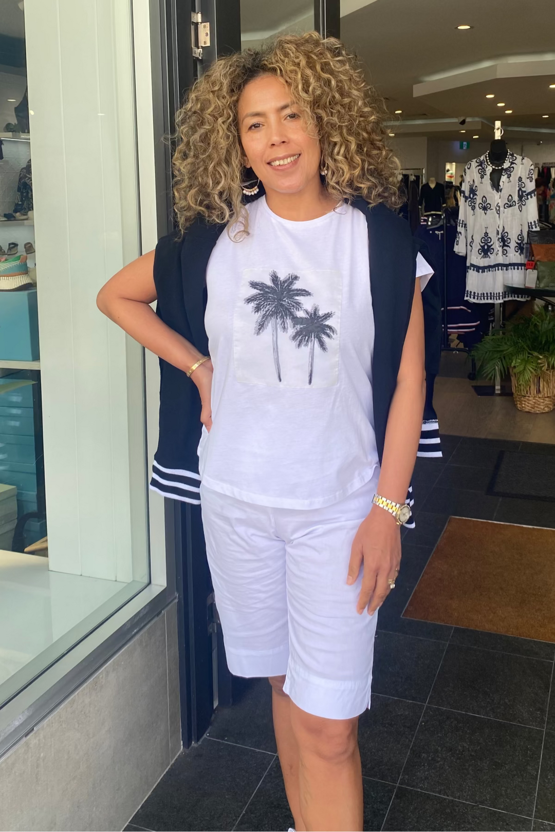 Woman standing in a store wearing a white t-shirt with palm tree design and white shorts.