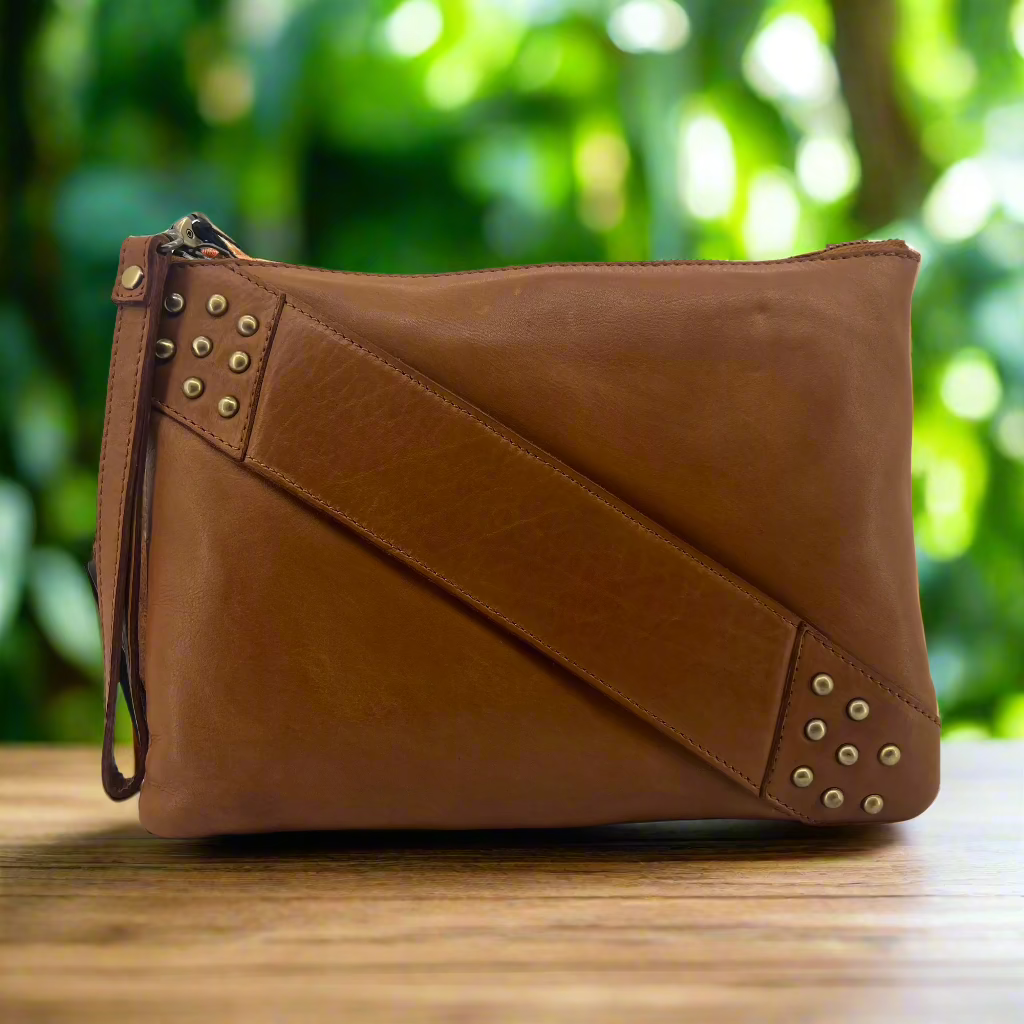 A tan-colored leather crossbody bag with an adjustable strap and studded details, displayed against a lush green background.