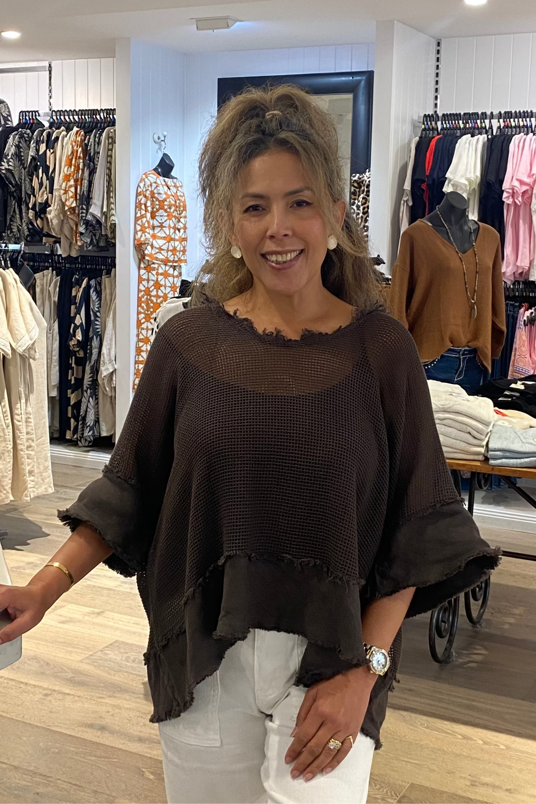 Woman standing in a clothing store wearing a brown top and white pants.