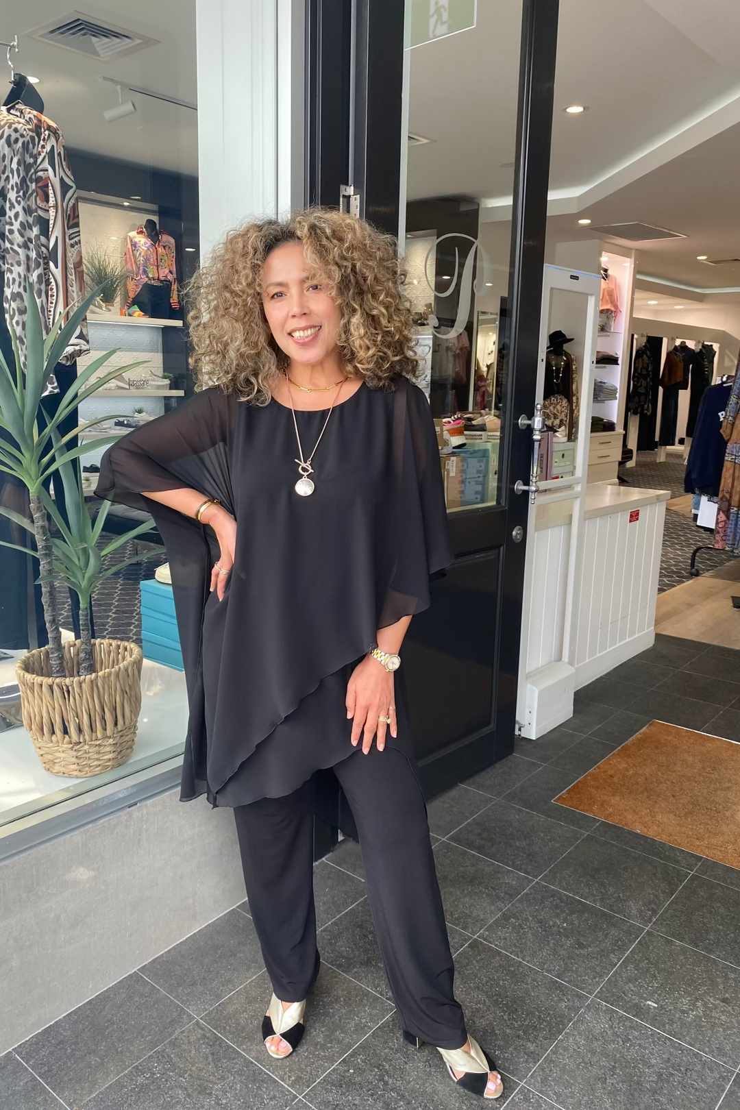 Woman in a black outfit standing outside a store with clothing displayed inside.