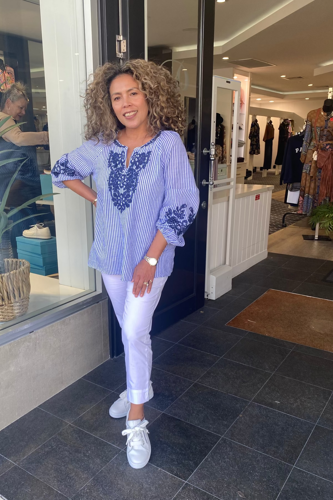 Woman in a store wearing a blue embroidered top and white pants.