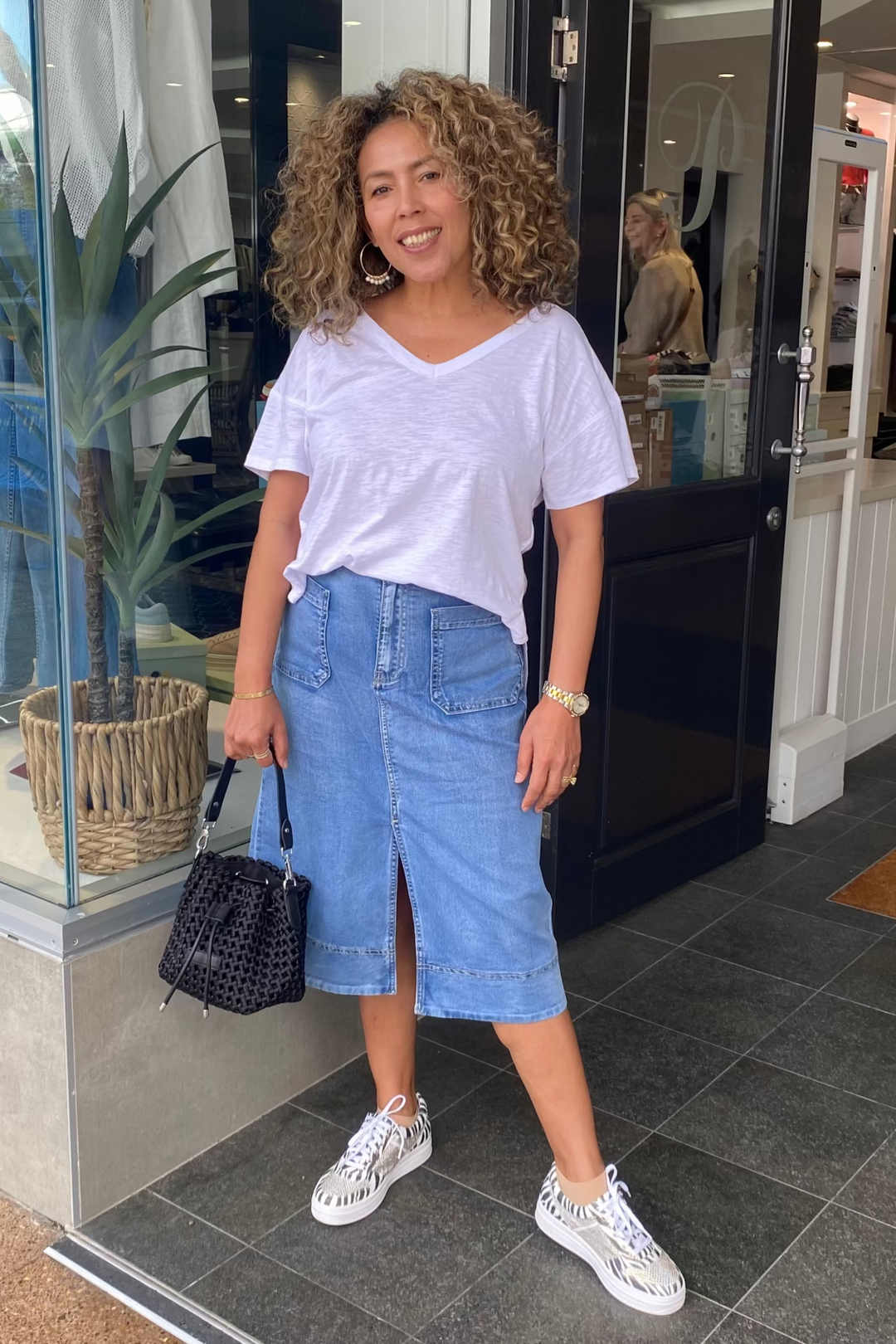 Woman standing outside a store wearing a white top and blue skirt.