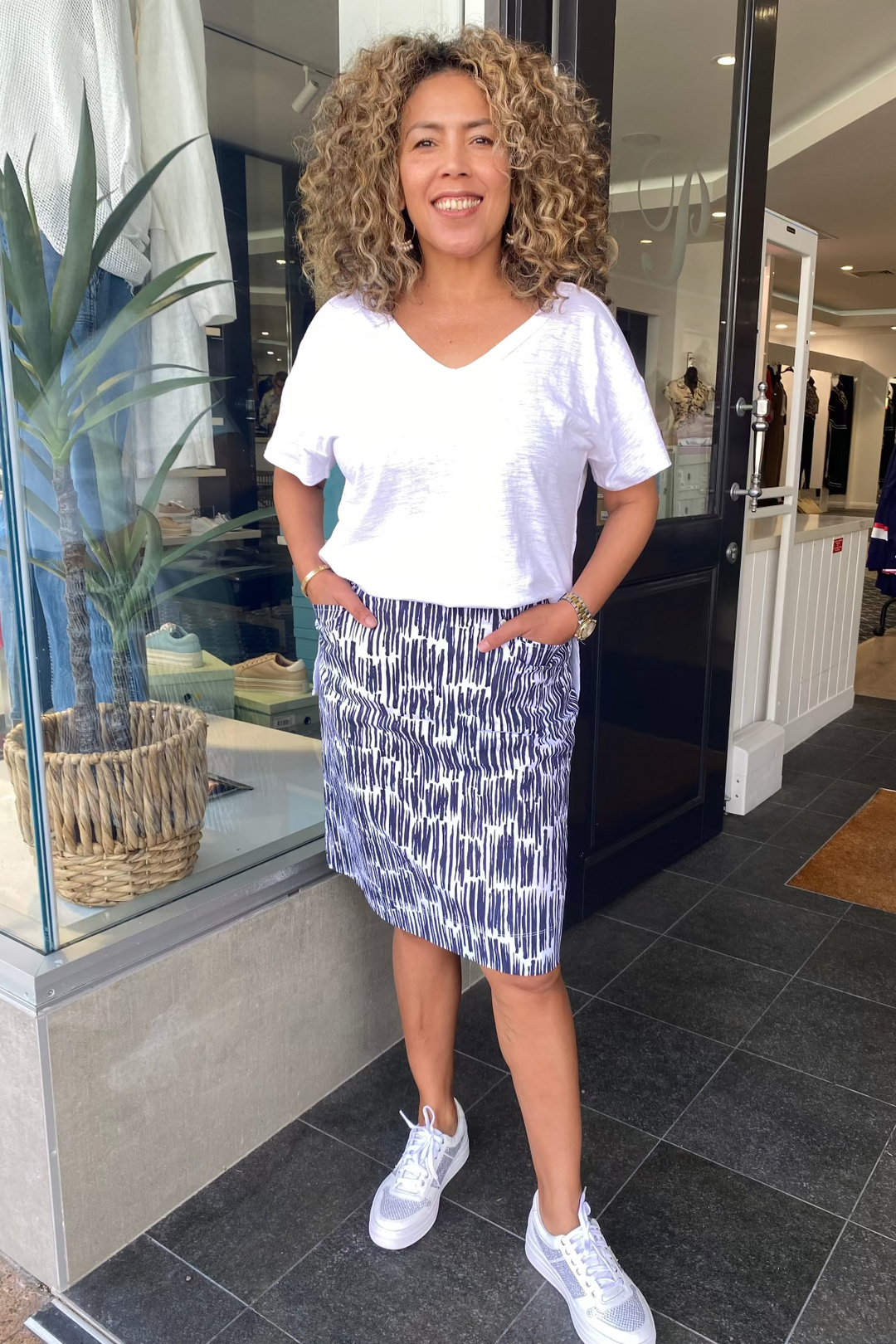 Woman standing in a store entrance wearing a white t-shirt and blue patterned skirt.