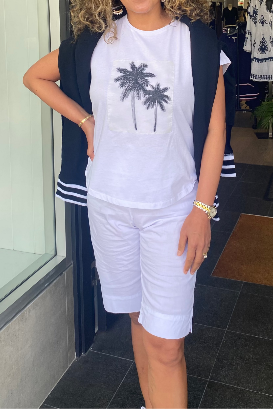 Woman standing in a store wearing a white t-shirt with palm tree design and white shorts.