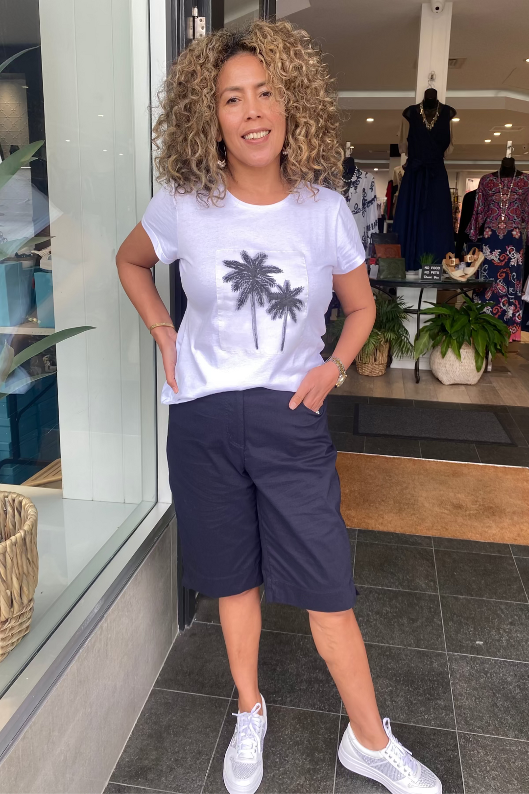 Woman wearing a white t-shirt with palm tree design and navy shorts standing in front of a store entrance.
