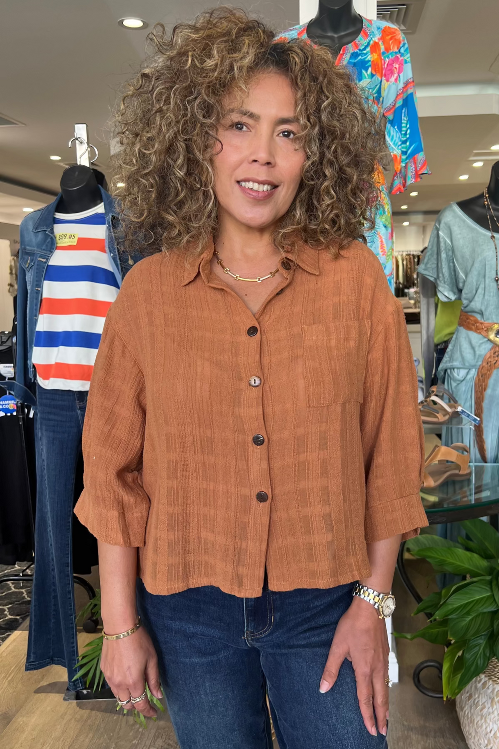 Woman standing in a clothing store wearing a brown shirt and blue jeans, surrounded by mannequins and clothing.