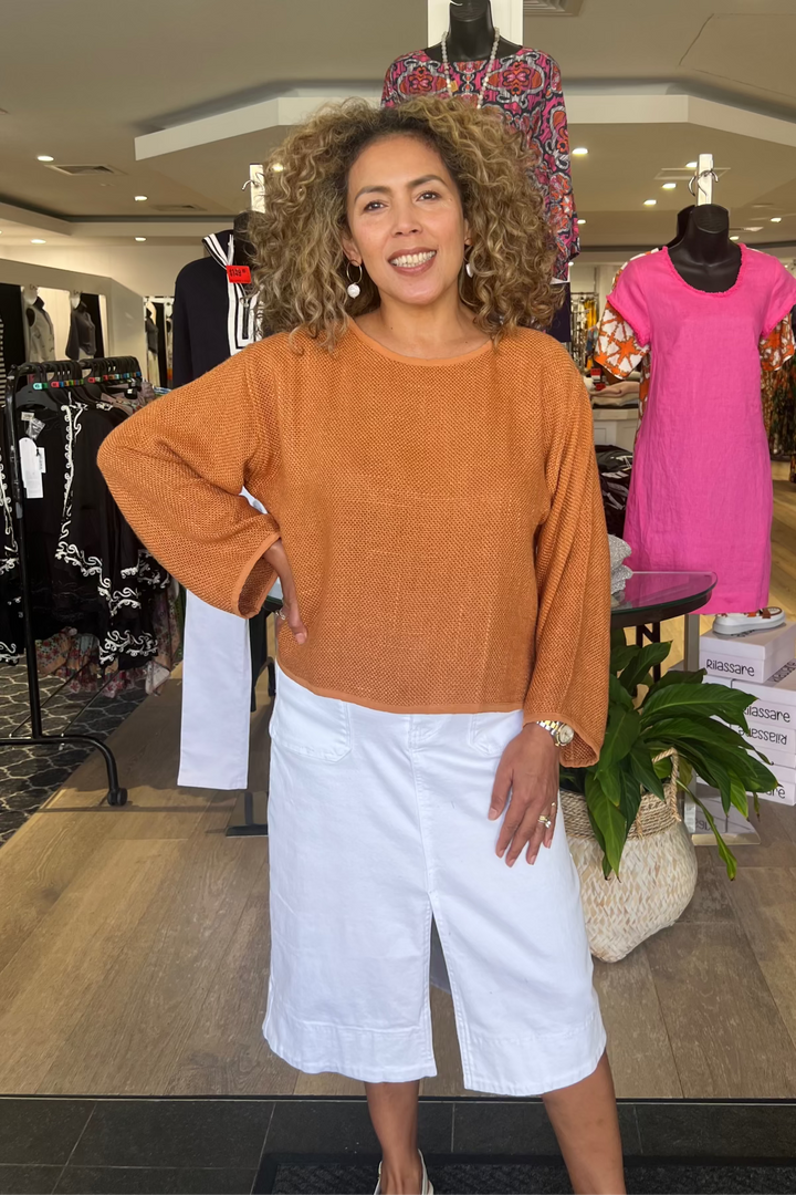 Woman posing in a clothing store wearing an orange sweater and white pants.