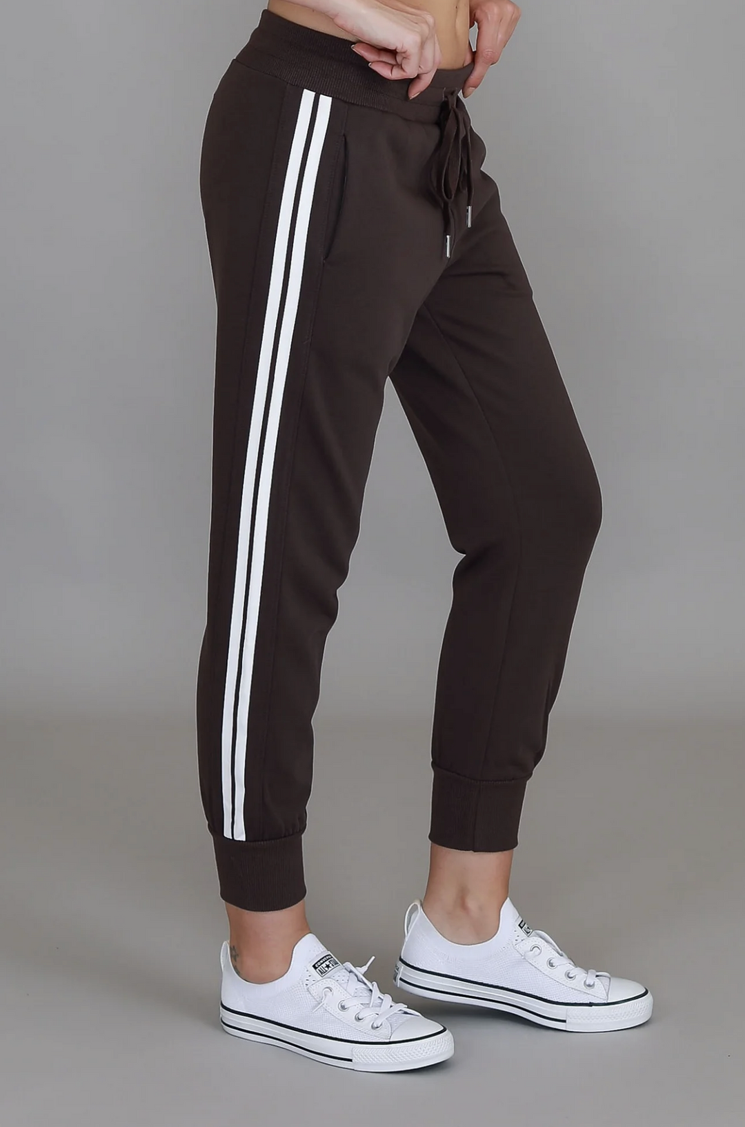 Brown jogger pants with white stripes worn by a person on a gray background