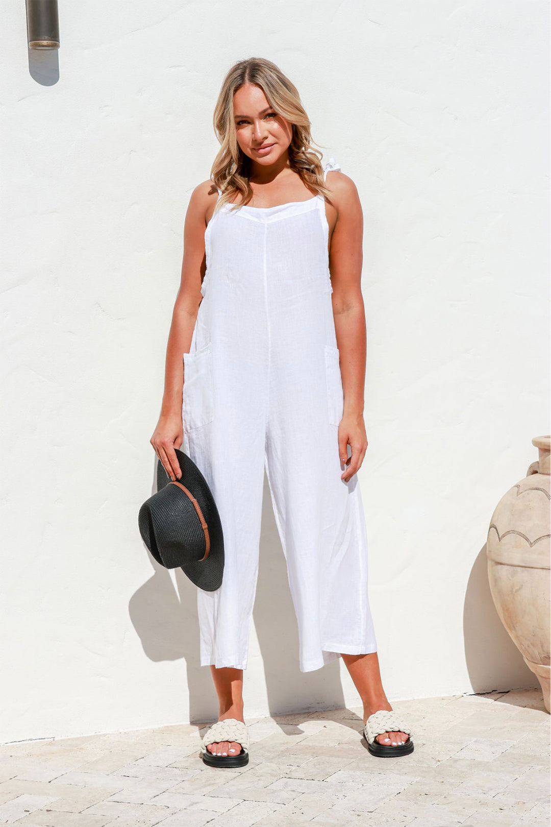 Full length photo of A lady standing in a white linen all in one jumpsuit with her hands in front pockets, the jumpsuit has wide 7/8 length legs with thin shoulder straps , the lady is also wearing a black sun hat, the linen jumpsuit is by The Italian cartel and Sold by Pizazz Boutique Nelson Bay NSW Australia