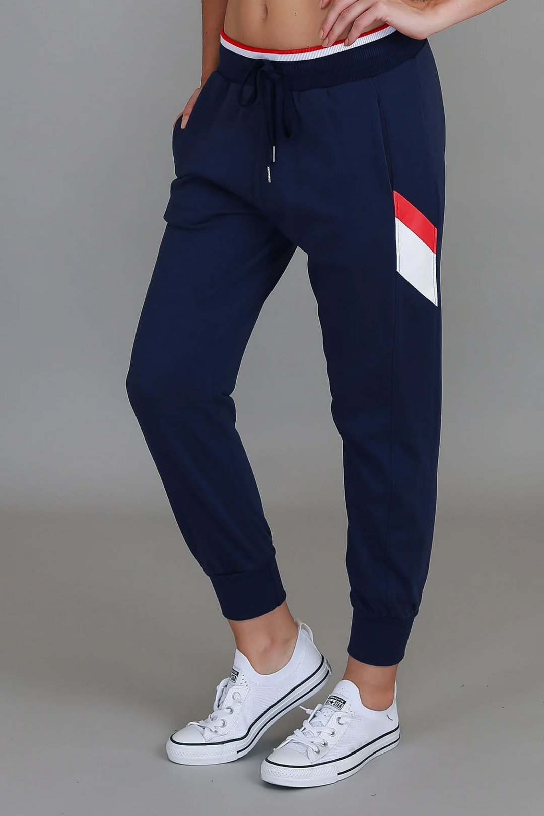 Navy jogger pants with red and white stripe worn by a person on a gray background