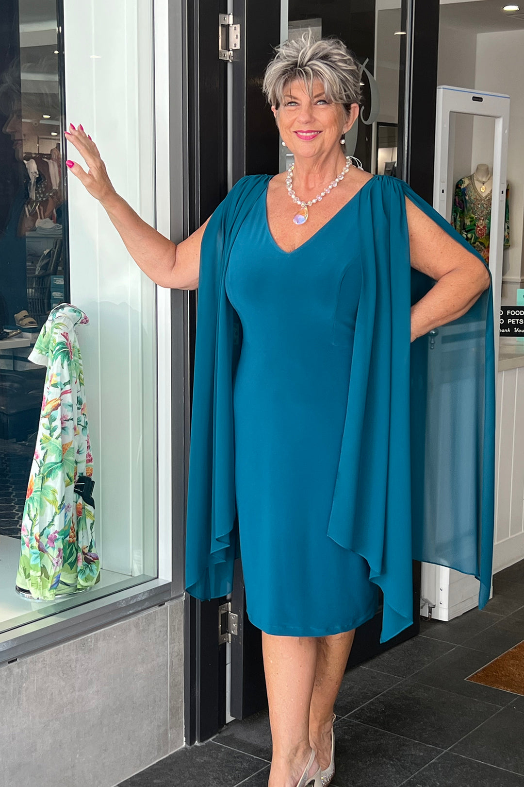 Lady wearing a blue evening gown, knee length with draping sleeves from Pizazz Boutique Nelson Bay